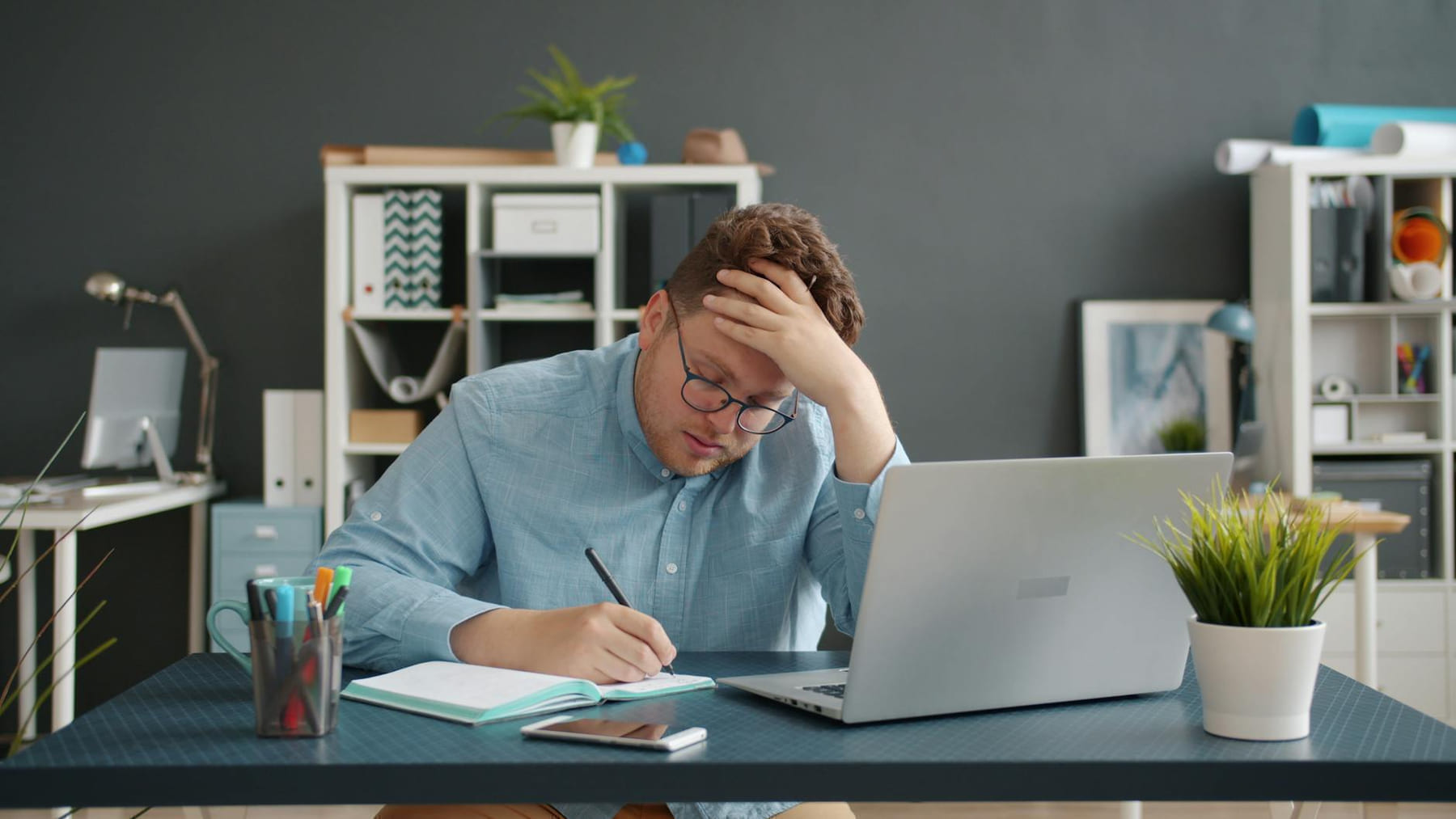 Man in office writes notes by laptop, looks stressed. Modern workspace. on The Digital Hustler