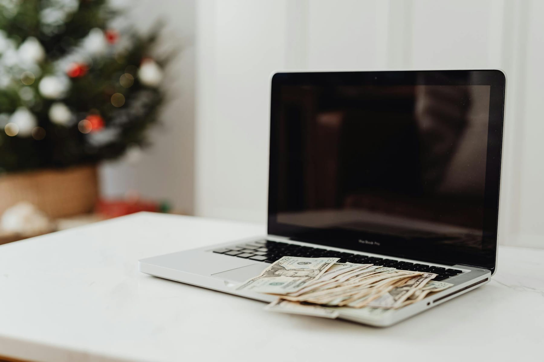 A laptop on a desk covered with cash, featuring a Christmas tree in the background. on The Digital Hustler